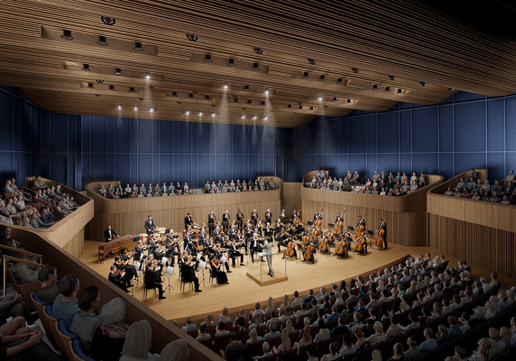 An orchestra performs on a large wooden stage in a modern concert hall filled with audience members on all sides.