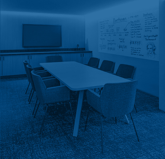Conference room with a long wooden table surrounded by yellow and brown chairs. A wall-mounted screen and a whiteboard with handwritten notes are visible.